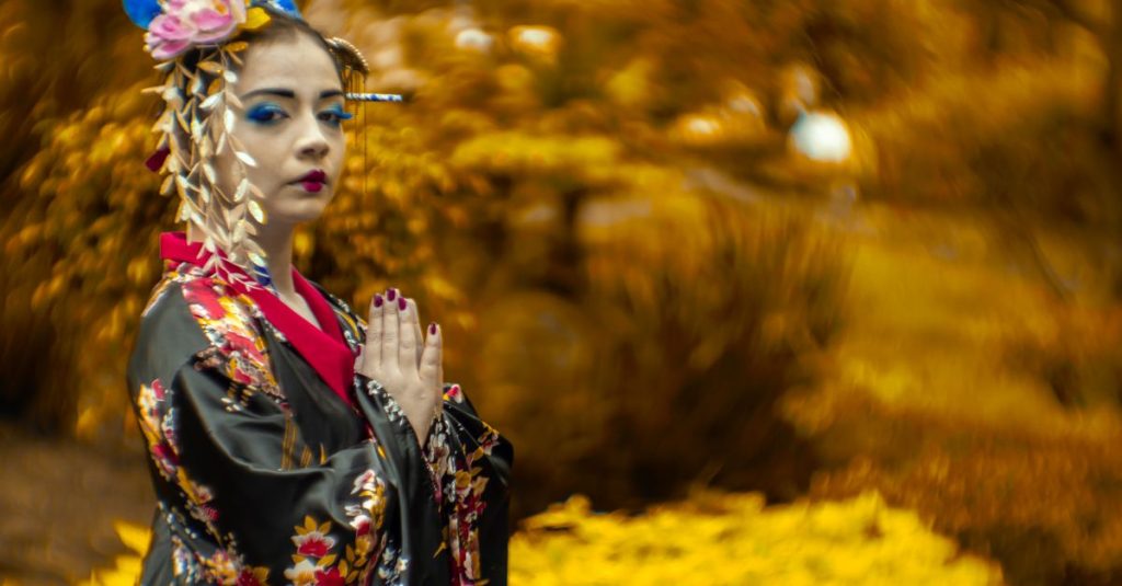 Femme maquillée et vêtue d'un kimono noir à fleurs, mains jointes, devant un feuillage doré