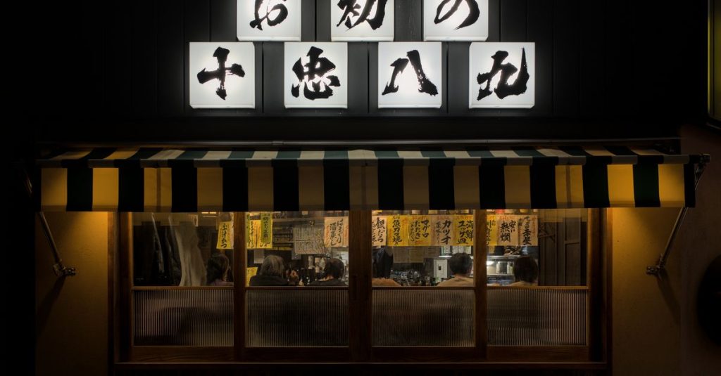 Façade éclairée de nuit d'un restaurant japonais, avec des lanternes portant des caractères japonais (l'image ne montre pas de tableau de hiragana)