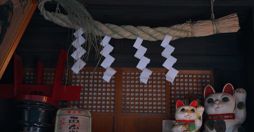 Corde sacrée en paille ornée de papiers en zigzag devant un petit sanctuaire, avec deux chats porte-bonheur