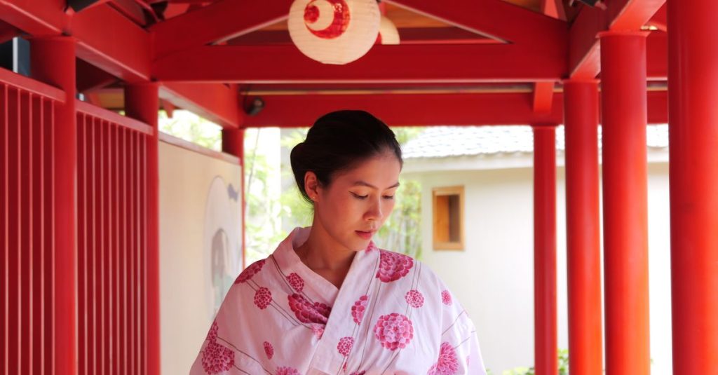 Femme en yukata blanc à motifs de fleurs roses, sous une galerie aux piliers rouges